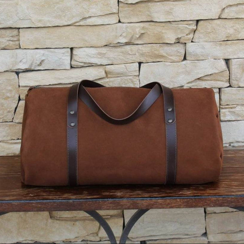 Brown duffel bag with leather straps on a wooden surface against a stone wall background