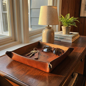 Brown leather tray on a wooden surface with sunglasses, keys, and a lamp in the background.